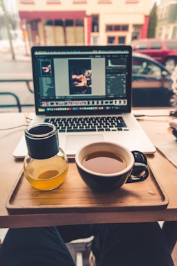 A cozy coffee shop setup with a laptop displaying photo editing software, placed on a table alongside a cup of tea and a small bottle of orange juice. The background shows a window view of a street with cars parked.
