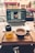 A cozy coffee shop setup with a laptop displaying photo editing software, placed on a table alongside a cup of tea and a small bottle of orange juice. The background shows a window view of a street with cars parked.