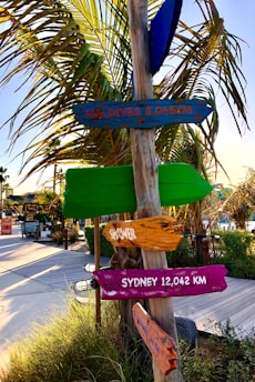 A colorful signpost stands next to a wooden pathway surrounded by greenery and palm trees. The signs point in various directions, indicating distances to distant locations like Maldives and Sydney.