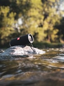 A portable speaker rests on a rock surrounded by flowing water, set against a backdrop of blurred green foliage. Sunlight filters through the trees, creating a serene and natural atmosphere.