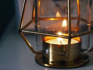 A lit candle is placed inside a decorative metallic frame with glass sides. The candlelight emits a warm glow that reflects softly on the surrounding surfaces, creating a cozy and intimate atmosphere.