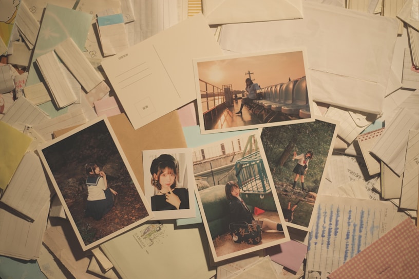 A collection of photographs laid out over a cluttered background of papers and envelopes. The images depict various scenes including a person sitting on outdoor seats at sunset, a person posing near a fence, and another in a school uniform in a wooded area. Some images have a nostalgic and candid feel.