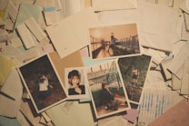 A collection of photographs laid out over a cluttered background of papers and envelopes. The images depict various scenes including a person sitting on outdoor seats at sunset, a person posing near a fence, and another in a school uniform in a wooded area. Some images have a nostalgic and candid feel.
