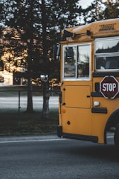 A yellow school bus is parked on the side of a road next to a chain-link fence. The bus has a stop sign extended and there are trees in the background, casting shadows on the ground.