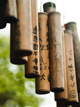 A collection of bamboo wind chimes with black calligraphy hanging outdoors, with a soft-focus background of greenery creating a natural and tranquil setting.
