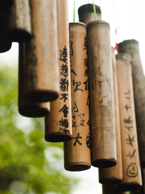 A collection of bamboo wind chimes with black calligraphy hanging outdoors, with a soft-focus background of greenery creating a natural and tranquil setting.