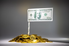 A meticulously arranged pile of shiny gold coins is topped by a rolled paper acting as a pole that holds a one hundred dollar bill. The setup resembles a flag mounted on a mound of wealth.