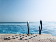 A tranquil swimming pool with clear blue water extends towards the horizon, meeting with the sky. Stainless steel pool ladders are prominently visible in the foreground, casting shadows on the wooden deck. Seagulls are seen flying in the distant sky, suggesting an open, coastal location.