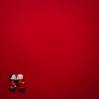 Two small figurines, styled in a traditional Chinese attire, stand close to each other against a vibrant red background. The figurines appear to be sharing a moment of affection, with one seemingly kissing the other's cheek.