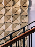 A geometric wall pattern with a 3D effect featuring light brown and beige tones. The pattern consists of interlocking diamond shapes with a shadow effect, creating depth. In the foreground, there is a staircase railing with dark metal bars and a wooden handrail.