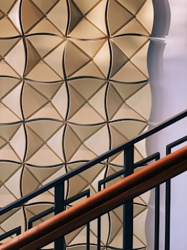 A geometric wall pattern with a 3D effect featuring light brown and beige tones. The pattern consists of interlocking diamond shapes with a shadow effect, creating depth. In the foreground, there is a staircase railing with dark metal bars and a wooden handrail.