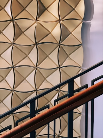 A geometric wall pattern with a 3D effect featuring light brown and beige tones. The pattern consists of interlocking diamond shapes with a shadow effect, creating depth. In the foreground, there is a staircase railing with dark metal bars and a wooden handrail.