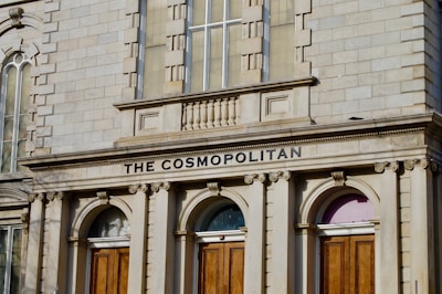 An ornate building facade made of light-colored stone features arched windows and wooden doors. The inscription 'THE COSMOPOLITAN' is prominently displayed above the doors.