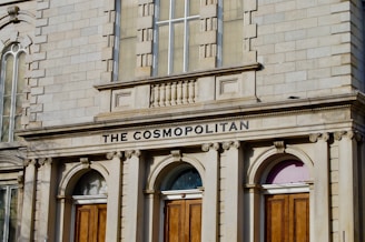 An ornate building facade made of light-colored stone features arched windows and wooden doors. The inscription 'THE COSMOPOLITAN' is prominently displayed above the doors.