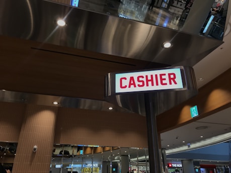An indoor setting with a large metallic structure and recessed lighting is prominently featured. A sign with the word 'CASHIER' in bold red letters is attached to a pole, drawing attention in the foreground. The surrounding area includes mirrored surfaces and reflects a spacious, possibly commercial environment.