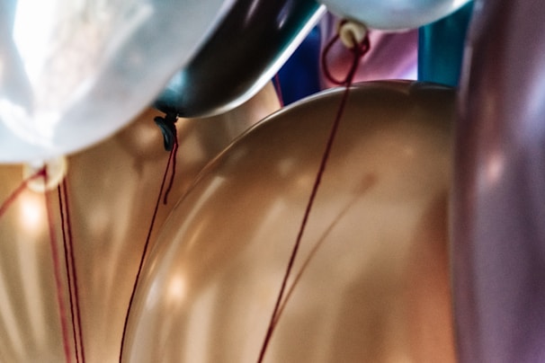 An arrangement of several balloons in close perspective, featuring a variety of colors including gold, silver, and hints of blue and pink. Some balloons are visible in the foreground while others are layered behind, with strings attached to each, adding dimension to the image.