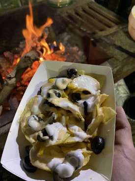 A serving of nachos topped with cheese sauce, sour cream, black olives, and corn. The dish is held close to a campfire, with flames visibly flickering in the background, creating a warm and cozy atmosphere.