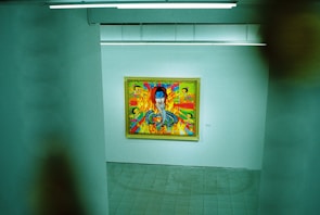 An art gallery features a brightly colored painting on a white wall. The artwork includes a vivid blue face surrounded by dynamic colors and patterns, with smaller figures or elements integrated into the scene.