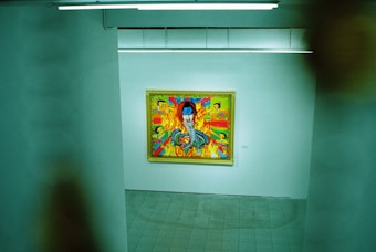 An art gallery features a brightly colored painting on a white wall. The artwork includes a vivid blue face surrounded by dynamic colors and patterns, with smaller figures or elements integrated into the scene.