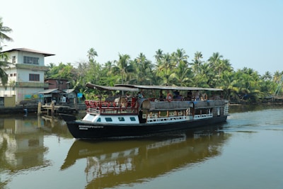 A houseboat navigates through a calm waterway surrounded by lush greenery and palm trees. There is a small pier with a few buildings nearby, reflecting the serene and tropical atmosphere.