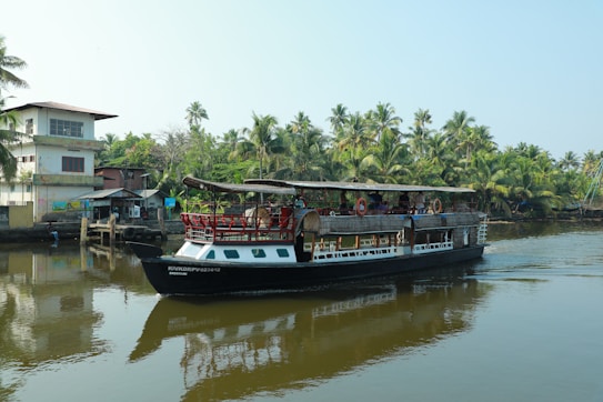 A houseboat navigates through a calm waterway surrounded by lush greenery and palm trees. There is a small pier with a few buildings nearby, reflecting the serene and tropical atmosphere.