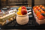An assortment of desserts is displayed, including a layered cake with cream and chocolate decorations, a white mug with fresh mango pieces and large strawberries on top, and small square containers of yogurt topped with strawberries and blueberries.