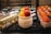 An assortment of desserts is displayed, including a layered cake with cream and chocolate decorations, a white mug with fresh mango pieces and large strawberries on top, and small square containers of yogurt topped with strawberries and blueberries.