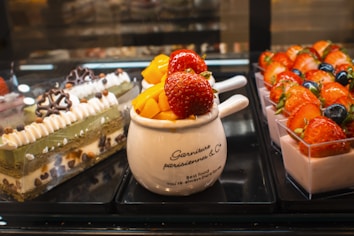 An assortment of desserts is displayed, including a layered cake with cream and chocolate decorations, a white mug with fresh mango pieces and large strawberries on top, and small square containers of yogurt topped with strawberries and blueberries.