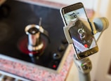 A smartphone is mounted on a gimbal stand, recording a video of a stovetop espresso maker placed on an active induction cooktop. The phone screen displays the live video capture of the brewing coffee pot, highlighted by the phone’s red recording button.
