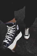 A pair of black high-top sneakers with white laces is worn by a person, partially visible from the knee down, standing on a dark surface. The shoes have a distinct round logo on the side, featuring a white star. The sneakers have thick white soles with a gum sole underneath, which is slightly visible.
