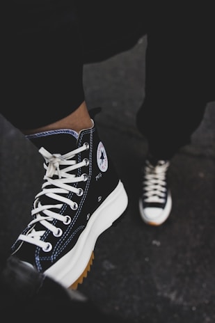 A pair of black high-top sneakers with white laces is worn by a person, partially visible from the knee down, standing on a dark surface. The shoes have a distinct round logo on the side, featuring a white star. The sneakers have thick white soles with a gum sole underneath, which is slightly visible.