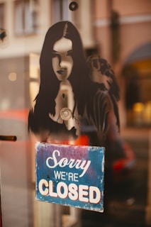 A stencil-style artwork of a woman with long hair is featured on a reflective glass surface. Below it, a blue and white sign reads 'Sorry We're Closed.' The background is slightly blurred, showing parts of a street scene.