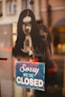 A stencil-style artwork of a woman with long hair is featured on a reflective glass surface. Below it, a blue and white sign reads 'Sorry We're Closed.' The background is slightly blurred, showing parts of a street scene.