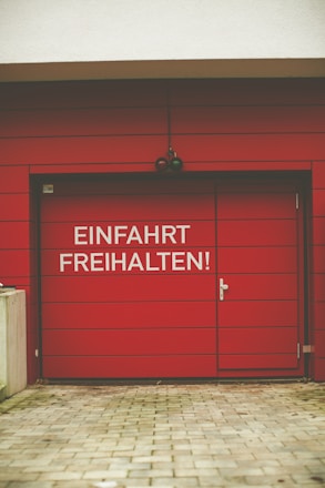 A red garage door with horizontal panels is prominent, featuring a white sign that reads 'Einfahrt Freihalten!' possibly indicating 'Keep Driveway Clear' in German. The door has a handle on the right side and is part of a building with a light-colored upper wall. The foreground consists of a paved stone pathway leading to the door.