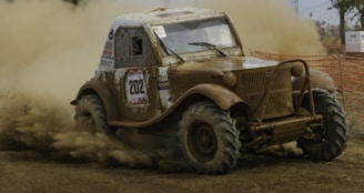 An off-road vehicle is navigating a muddy track, kicking up a significant amount of dust and mud. The vehicle is a rugged, vintage design with a number 202 decal on the side. The environment appears to be a competitive race setting with orange safety netting along the track and spectators in the background.