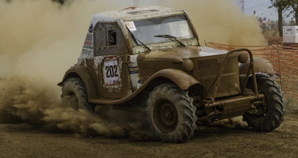 An off-road vehicle is navigating a muddy track, kicking up a significant amount of dust and mud. The vehicle is a rugged, vintage design with a number 202 decal on the side. The environment appears to be a competitive race setting with orange safety netting along the track and spectators in the background.