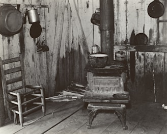 A rustic interior scene featuring a weathered wooden chair and an old cast iron stove. Several cooking pots and a kettle are hanging on the walls, which are made of rough wooden planks. Piles of chopped wood are stacked near the stove.