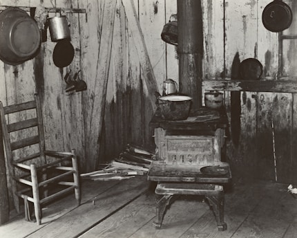 A rustic interior scene featuring a weathered wooden chair and an old cast iron stove. Several cooking pots and a kettle are hanging on the walls, which are made of rough wooden planks. Piles of chopped wood are stacked near the stove.