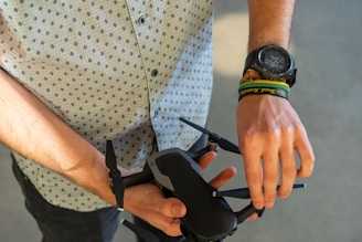 A person wearing a patterned shirt and a large wristwatch is holding a small black drone. Several colorful wristbands are also visible on one wrist. The drone is compact, with four propellers visible, and is being held in both hands.