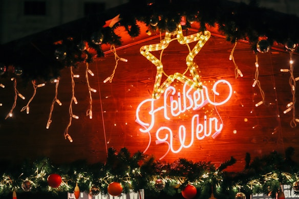 A glowing neon sign displays a stylized star and cursive text reading 'Heißer Wein' on a wooden background. The sign is surrounded by festive decorations, including hanging string lights and garlands with red ornaments.