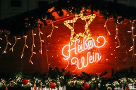 A glowing neon sign displays a stylized star and cursive text reading 'Heißer Wein' on a wooden background. The sign is surrounded by festive decorations, including hanging string lights and garlands with red ornaments.