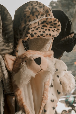 A variety of plush hats designed to resemble animal heads are displayed. The central hat is styled like a cheetah, with spotted fur and a black nose. Other hats feature different animal prints and textures.
