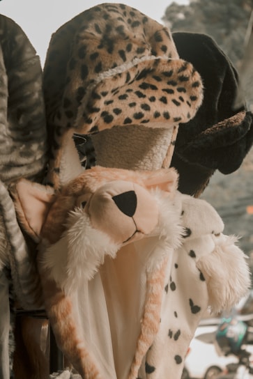 A variety of plush hats designed to resemble animal heads are displayed. The central hat is styled like a cheetah, with spotted fur and a black nose. Other hats feature different animal prints and textures.