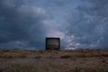 A vintage television set sits on a barren, grassy field under a dramatic sky with dark and moody clouds. The horizon is low, making the sky dominate the scene, with hints of purple and blue in the clouds suggesting an approaching or receding storm.