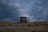 A vintage television set sits on a barren, grassy field under a dramatic sky with dark and moody clouds. The horizon is low, making the sky dominate the scene, with hints of purple and blue in the clouds suggesting an approaching or receding storm.