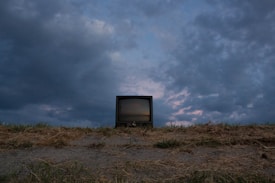 A vintage television set sits on a barren, grassy field under a dramatic sky with dark and moody clouds. The horizon is low, making the sky dominate the scene, with hints of purple and blue in the clouds suggesting an approaching or receding storm.