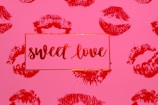 A pink background features multiple red lipstick kiss marks. In the center, a rectangular box with a thin red border contains the phrase 'sweet love' written in elegant, flowing red script.