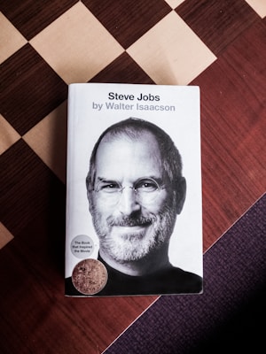 A hardcover book titled 'Steve Jobs' by Walter Isaacson is placed on a checkered surface. The cover features a black and white portrait of a man with glasses and a beard. Two stickers are attached to the cover, one indicating the book inspired a movie.