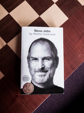 A hardcover book titled 'Steve Jobs' by Walter Isaacson is placed on a checkered surface. The cover features a black and white portrait of a man with glasses and a beard. Two stickers are attached to the cover, one indicating the book inspired a movie.