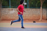 A person in a red t-shirt and blue jeans is running on an outdoor court with a background of a stone wall and fencing. There is a single black shoe on the ground behind the person, and trees are visible in the background.